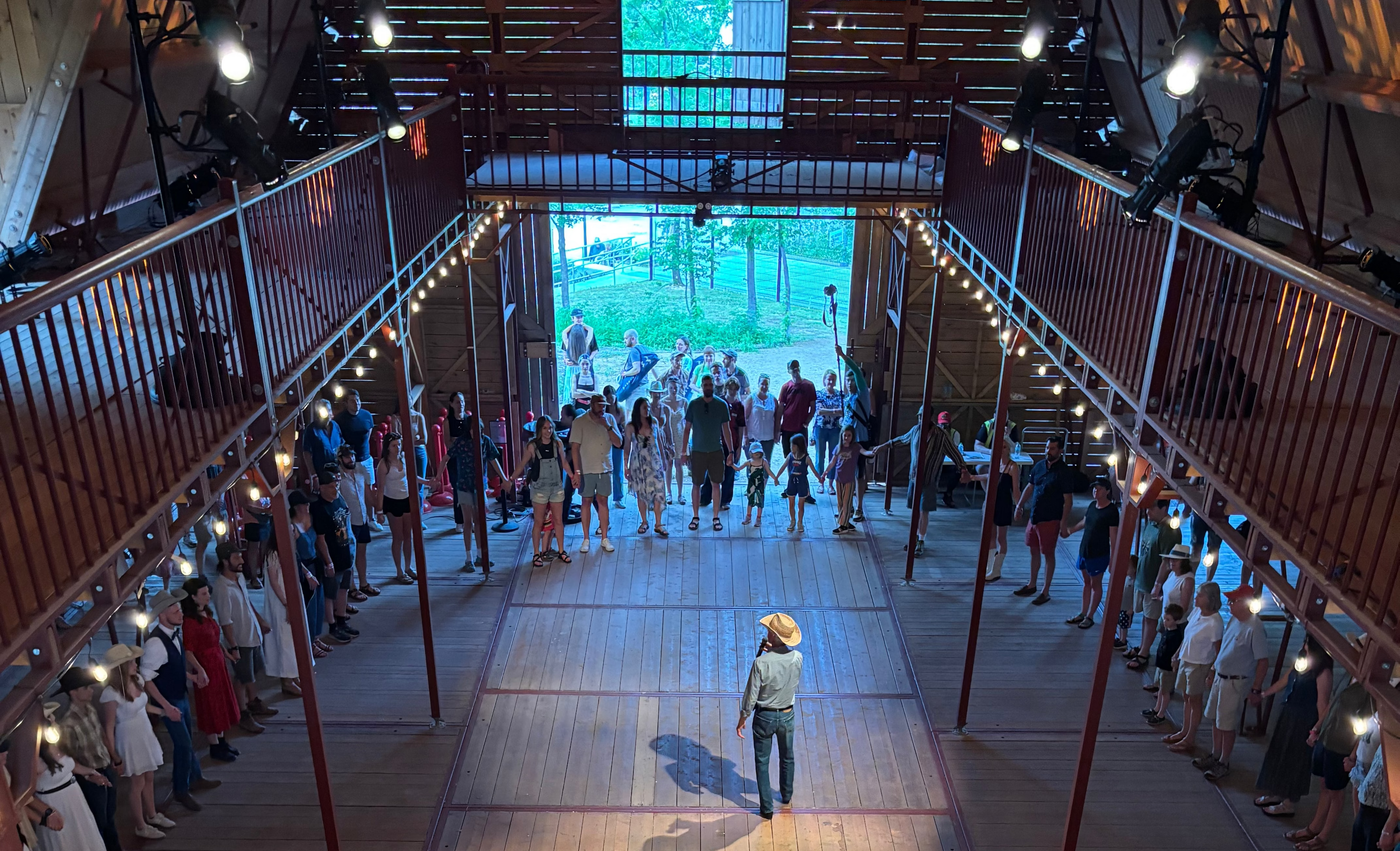 Group of people holding hands in a large indoor wooden hall with a stage and audience.